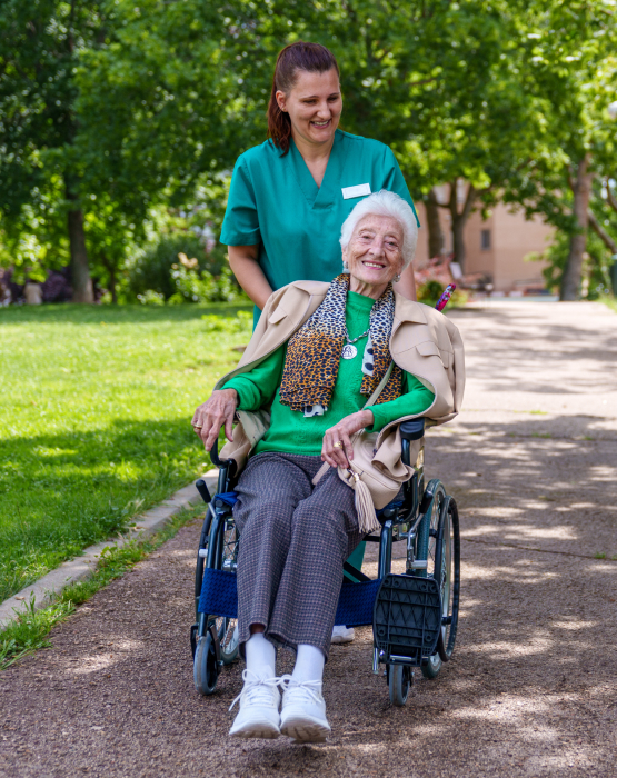 Smiling care worker pushes elderly woman in a wheelchair through a park, providing outdoor elderly care